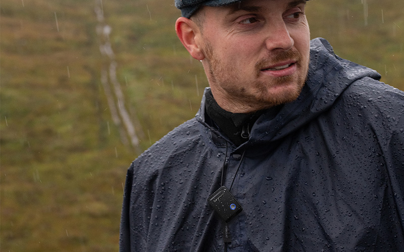 A man in a raincoat with a wireless microphone clipped to his jacket, standing in the rain.